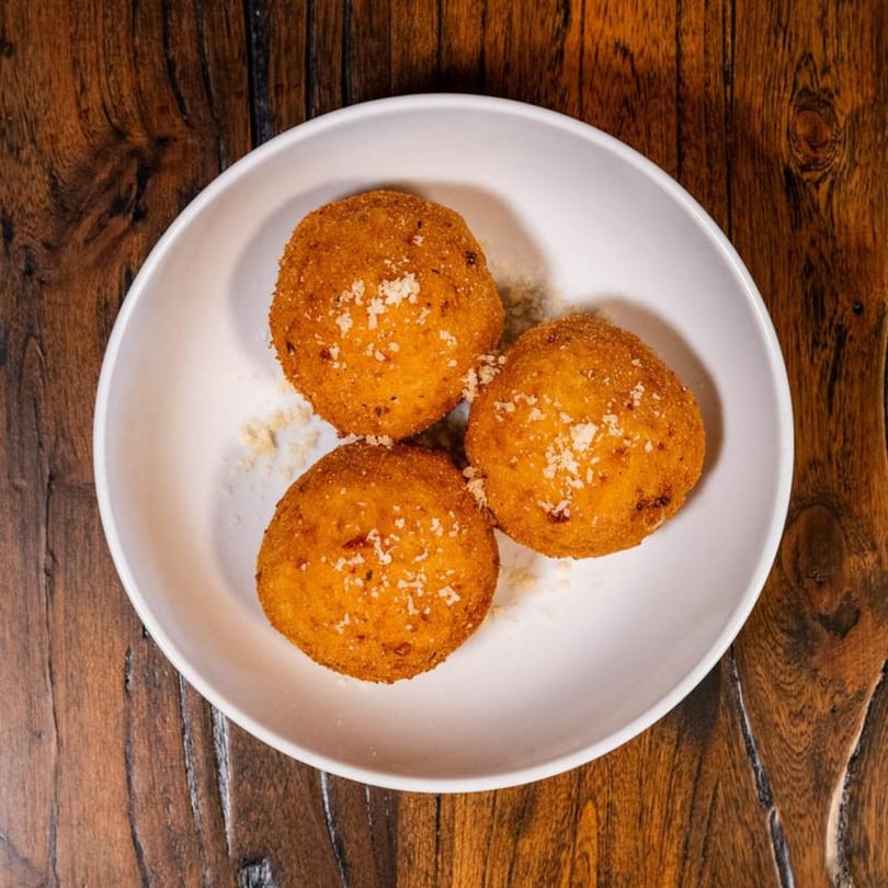 Three golden-brown arancini balls, sprinkled with parmesan cheese, in a white bowl on a wooden table.