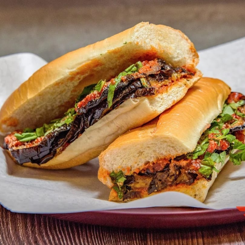 Two eggplant parmesan subs with red sauce and fresh greens on white paper, on a dark red plate.