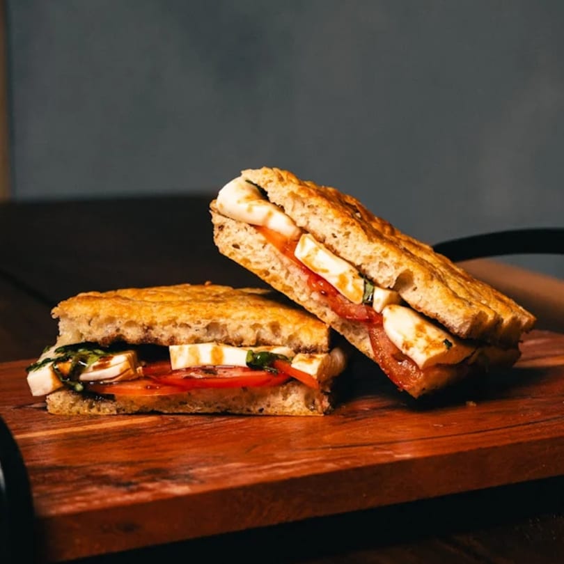 Two halves of a grilled panini sandwich with mozzarella, tomato, and basil on a wooden board.