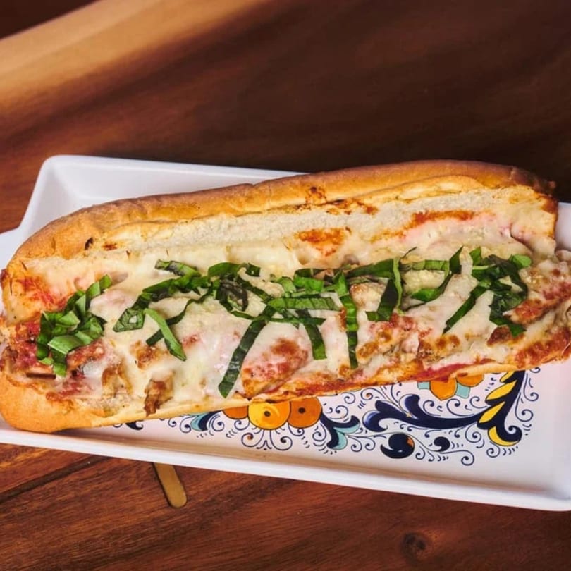 A chicken parmesan sub sandwich with melted cheese and fresh basil on a decorative white platter.