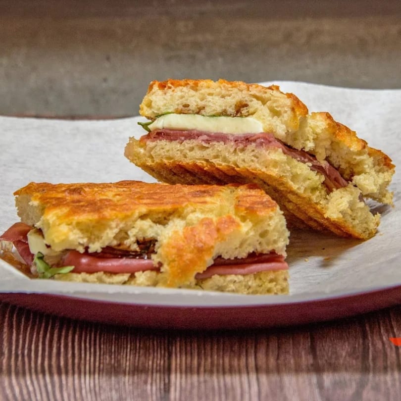 Two halves of a grilled panini sandwich with meat, cheese, and greens on a white plate.