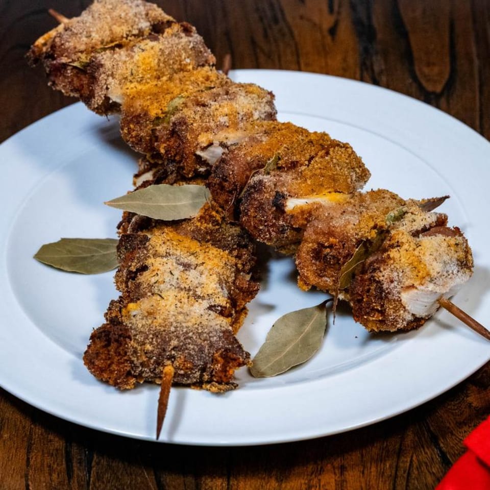 Breaded meat skewers with bay leaves on a white plate, on a wooden table.