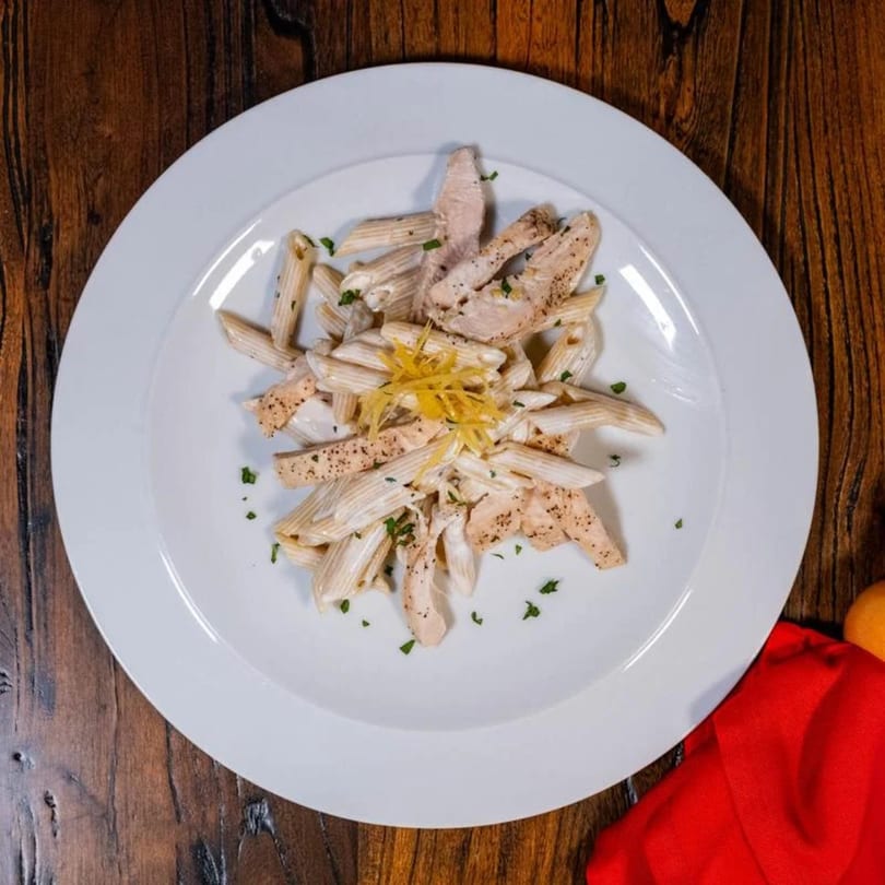 Creamy penne pasta with sliced chicken, black pepper, parsley, and lemon zest on a white plate.