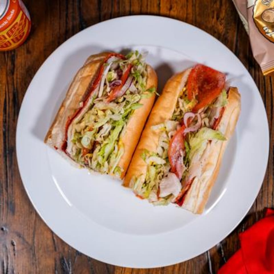 Two deli sandwiches with meat, lettuce, tomato, and onion on a white plate with a soda and chips.
